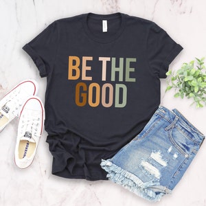 May include: A black t-shirt with the text "BE THE GOOD" in colorful letters. The letters are in shades of orange, brown, pink, and green.