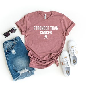 May include: A mauve colored t-shirt with the text "Stronger Than Cancer" and a white ribbon graphic. The t-shirt is laid out on a white background with a pair of denim shorts, sunglasses, and white sneakers.