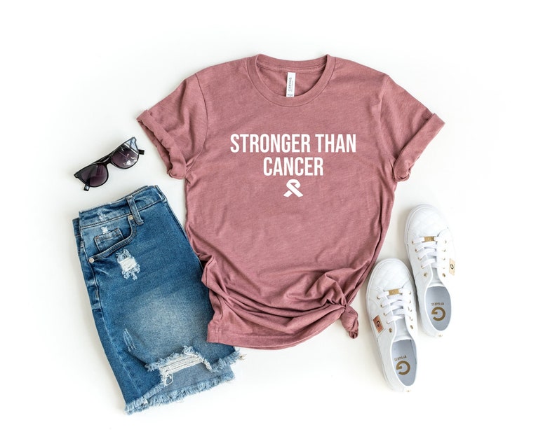 May include: A mauve colored t-shirt with the text "Stronger Than Cancer" and a white ribbon graphic. The t-shirt is laid out on a white background with a pair of denim shorts, sunglasses, and white sneakers.