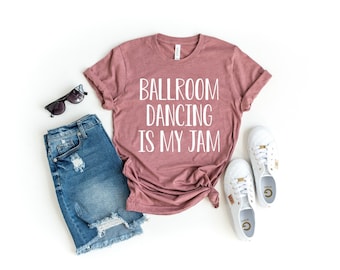 May include: A mauve t-shirt with the text "Ballroom Dancing is My Jam" in white lettering. The shirt is laid out on a white background with a pair of distressed denim shorts, sunglasses, and white sneakers.