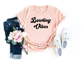 May include: A light pink t-shirt with the text "Bowling Vibes" in black retro-style lettering. The shirt is laid flat on a white background with a pair of white sneakers, blue jeans, and pink flowers.