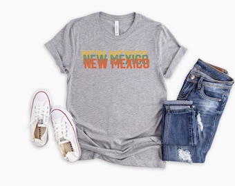 May include: A gray t-shirt with the text "NEW MEXICO" printed in yellow, orange, and green. The shirt is paired with blue jeans and white sneakers.