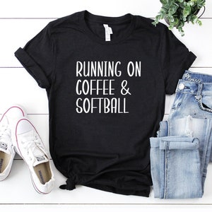 May include: Black t-shirt with the text "RUNNING ON COFFEE & SOFTBALL" in white. The shirt is paired with white sneakers and blue jeans. A small potted plant is visible in the background.