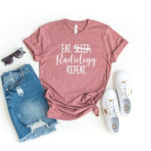 May include: A dusty rose t-shirt with the text "EAT. SLEEP. Radiology REPEAT." is paired with denim shorts, white sneakers, and sunglasses. The shirt is knotted at the waist, and the items are arranged on a white surface.