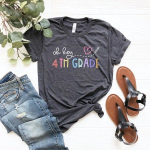Puede incluir: Una camiseta gris oscuro con el texto "oh hey 4th GRADE" en colores arcoíris. Un corazón con un lápiz dibujado a través de él está por encima del texto. La camiseta está colocada sobre una superficie blanca con un par de sandalias marrones y unos vaqueros azules.