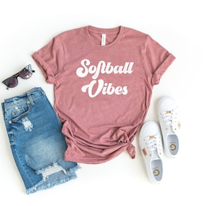 May include: A mauve t-shirt with the text "Softball Vibes" in white retro font. The shirt is laid out on a white surface with a pair of blue denim shorts, sunglasses, and white sneakers.