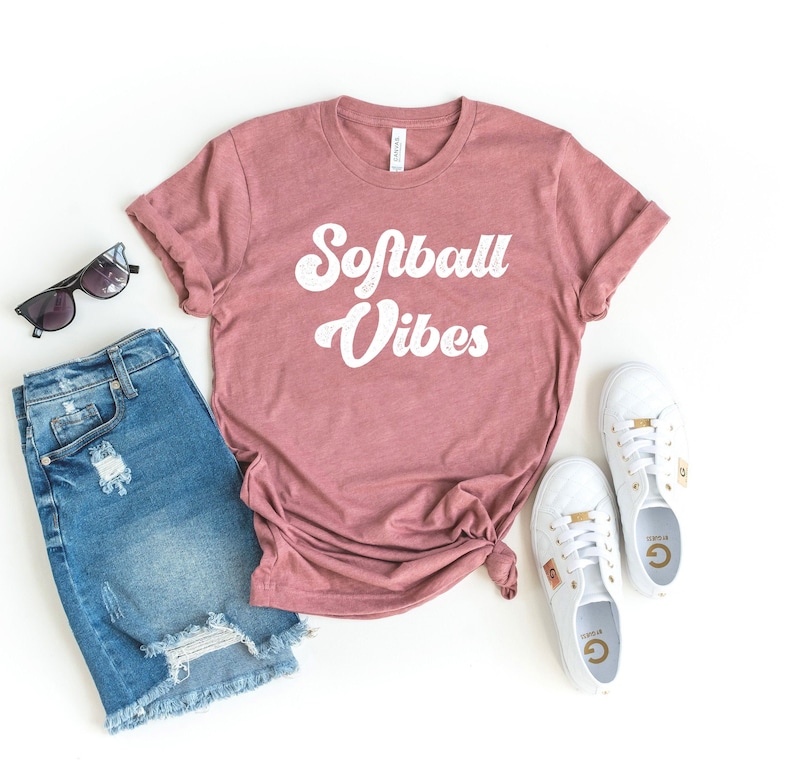 May include: A mauve t-shirt with the text "Softball Vibes" in white retro font. The shirt is laid out on a white surface with a pair of blue denim shorts, sunglasses, and white sneakers.