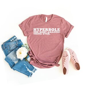 May include: A mauve t-shirt with the text "HYPERBOLE THE MOST AMAZING THING EVER." printed in white.