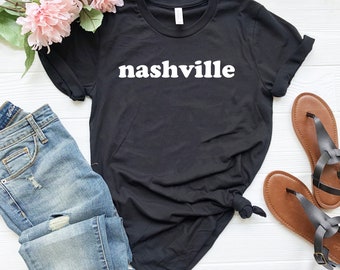 Puede incluir: Camiseta negra con texto blanco que dice "nashville" en una fuente retro. La camiseta se combina con jeans azules y sandalias marrones.