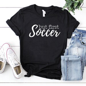 May include: Black t-shirt with white text that reads "but first soccer". The shirt is laying on a white wooden surface with a pair of white sneakers and blue jeans.