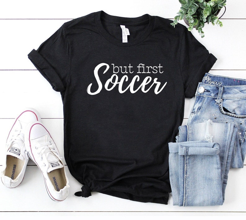 May include: Black t-shirt with white text that reads "but first soccer". The shirt is laying on a white wooden surface with a pair of white sneakers and blue jeans.