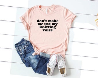 以下が含まれることがあります： 黒い文字で「don't make me use my knitting voice」と書かれたピンクのTシャツ