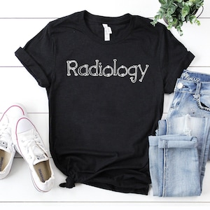 May include: Black t-shirt with the word "Radiology" written in white, with the letters made up of bone shapes. The shirt is laying on a white surface with a pair of white sneakers and a pair of blue jeans.