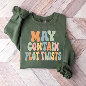 May include: An olive green crewneck sweatshirt featuring the text MAY CONTAIN PLOT TWISTS printed in a multi-colored, wavy font. The letters are colored in shades of orange, light blue, sage green, and soft pink against the dark green fabric.