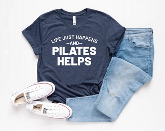 May include: A navy blue t-shirt with white text that reads "Life just happens -and- Pilates helps". The shirt is paired with blue jeans and white sneakers.