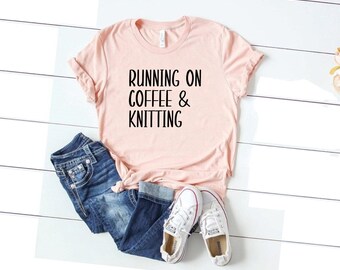 Puede incluir: Una camiseta rosa con el texto "RUNNING ON COFFEE & KNITTING" en letras negras. La camiseta está colocada sobre una superficie de madera blanca con un par de zapatillas blancas y unos vaqueros azules.