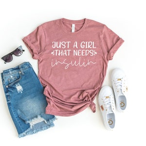 May include: A mauve t-shirt with the text "Just a girl that needs insulin" in white lettering. The shirt is laid out on a white surface with a pair of blue denim shorts, sunglasses, and white sneakers.