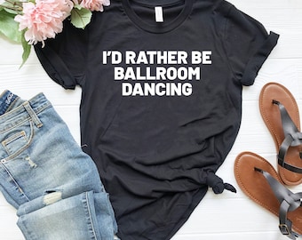 May include: Black t-shirt with white text that reads "I'd rather be ballroom dancing". The shirt is laid flat on a wooden surface with a pair of blue jeans and brown sandals.