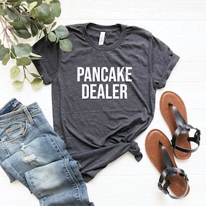 Pu&ograve; includere: Una maglietta grigia scura melange con la scritta "PANCAKE DEALER" stampata in bianco.