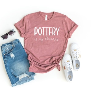 Może przedstawiać: Koszulka w kolorze malwy z napisem "POTTERY is my therapy" w białym kolorze. Koszulka leży na białym tle z niebieskimi dżinsowymi spodenkami, okularami przeciwsłonecznymi i białymi butami sportowymi.