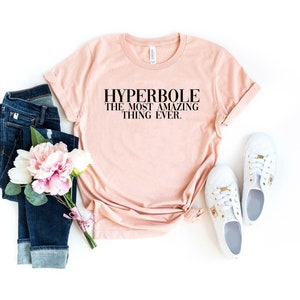 Puede incluir: Una camiseta rosa claro con el texto "HYPERBOLE THE MOST AMAZING THING EVER." impreso en negro.