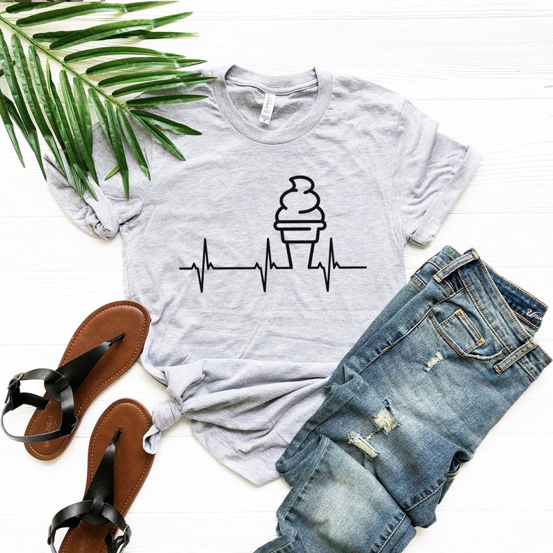 Peut inclure: Un t-shirt gris avec un motif de cornet de glace noir et une ligne de battement de c&oelig;ur. Le cornet de glace est au centre du t-shirt et la ligne de battement de c&oelig;ur est en dessous. Le t-shirt est pos&eacute; sur une surface blanche avec une paire de sandales marrons et un jean bleu.