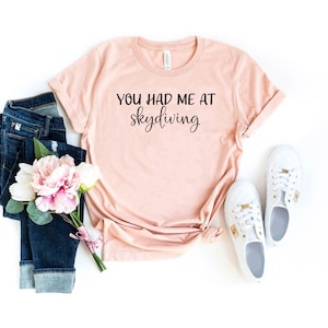 May include: A light pink t-shirt with the text "You had me at skydiving" in black lettering. The shirt is laid out on a white background with a pair of blue jeans, a bouquet of pink flowers, and two white sneakers.