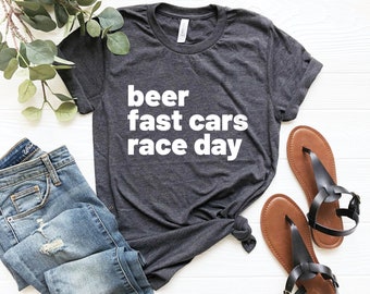 Może przedstawiać: Szary t-shirt z nadrukiem "beer fast cars race day" w kolorze białym na przodzie. Koszulka leży na białym tle z parą brązowych sandałów i niebieskimi dżinsami.