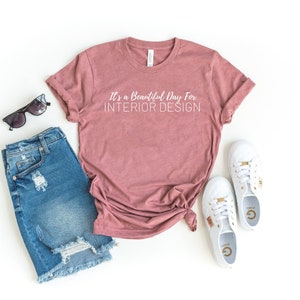 May include: A mauve t-shirt with the text "It's a Beautiful Day For INTERIOR DESIGN" in white. The t-shirt is laid out on a white surface with a pair of blue denim shorts, sunglasses, and white sneakers.