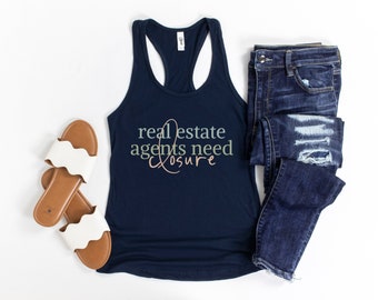 May include: A navy blue tank top with the text "real estate agents need closure" in green, pink, and white lettering. The tank top is paired with a pair of blue jeans and white sandals.
