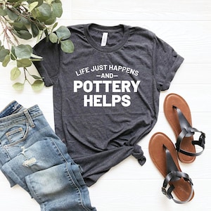 Puede incluir: Una camiseta gris oscuro con el texto "Life just happens -and- Pottery helps" en letras blancas.