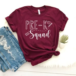 Peut inclure: T-shirt bordeaux avec un texte blanc qui dit "Pre-K Squad" avec un cœur, un crayon et un livre graphique.