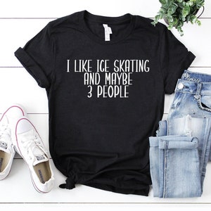 May include: Black t-shirt with the white text "I LIKE ICE SKATING AND MAYBE 3 PEOPLE." The shirt is paired with white sneakers and blue jeans. A small potted plant is visible in the background.