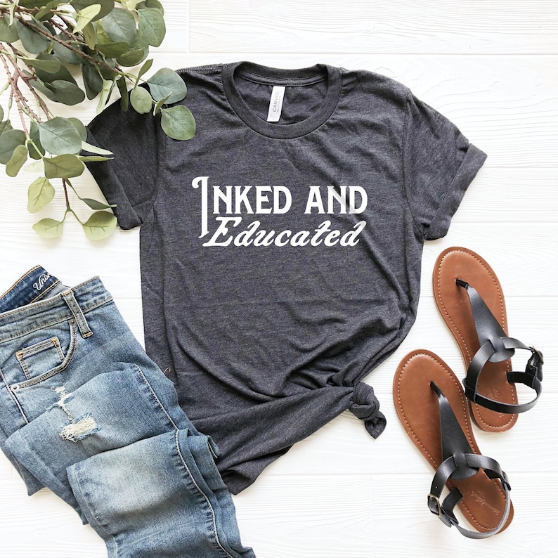 May include: A dark gray t-shirt with the text "INKED AND Educated" in white lettering. The shirt is laid out on a white wooden surface with a pair of brown sandals and blue jeans.