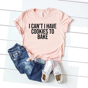 Puede incluir: Una camiseta rosa con el texto "I can't I have cookies to bake" en letras negras.