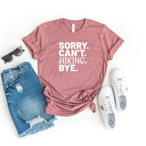 May include: A mauve t-shirt with the text "SORRY. CAN'T. HIKING. BYE." in white. The shirt is laid out on a white background with a pair of blue denim shorts, sunglasses, and white sneakers.