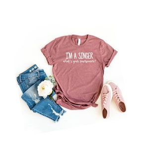 May include: A mauve t-shirt with the text "I'm a singer what's your superpower?" printed in white. The t-shirt is laid flat on a white background with a pair of light pink trainers, a pair of blue jeans, and a white flower.