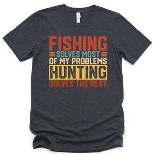 以下が含まれることがあります： ヴィンテージスタイルのグラフィックがプリントされたダークヘザーグレーのTシャツ。グラフィックには「Fishing solves most of my problems Hunting solves the rest」と書いてあります。