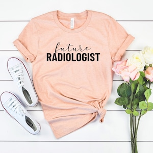 May include: A peach-colored t-shirt with the words "future RADIOLOGIST" printed in black. The shirt is styled with white sneakers and a bouquet of pink and white flowers. The shirt is knotted at the waist.