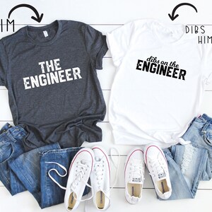 May include: Two t-shirts with text, jeans, and white sneakers. One dark gray t-shirt reads "THE ENGINEER" in white block letters. The other is white and reads "dibs on the ENGINEER" in black. Both shirts are paired with blue jeans and white sneakers.