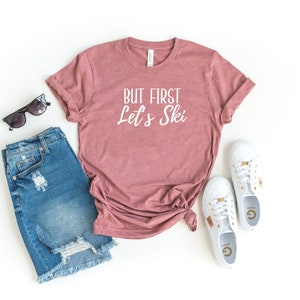 May include: A mauve t-shirt with the text "But First Let's Ski" in white lettering. The shirt is laid out on a white surface with a pair of blue denim shorts, sunglasses, and white trainers.