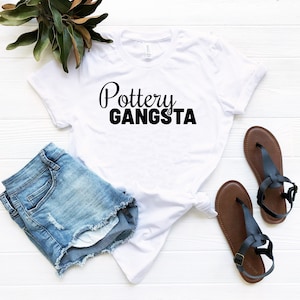 May include: White t-shirt with black text that reads "Pottery Gangsta". The shirt is lying on a white wooden surface with a pair of blue denim shorts and brown sandals.