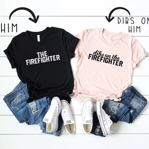 以下が含まれることがあります： テキストが書かれた2枚のTシャツ。黒いTシャツには「The Firefighter」と書いてあり、ピンクのTシャツには「Dibs on the Firefighter」と書いてあります。