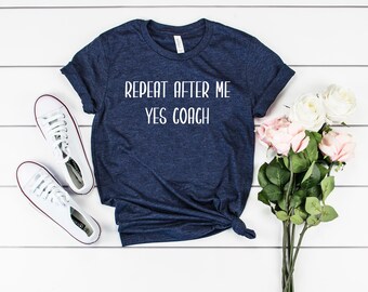 Puede incluir: Una camiseta azul marino con el texto "Repeat after me Yes Coach" en letras blancas. La camiseta está colocada sobre una superficie de madera blanca con un par de zapatillas blancas y un ramo de rosas rosas y blancas.