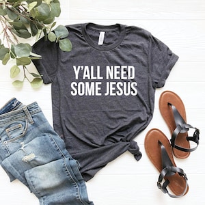 May include: A dark gray t-shirt with the text "Y'ALL NEED SOME JESUS" printed in white on the front. The shirt is laid out on a white surface with a pair of brown sandals and blue jeans.