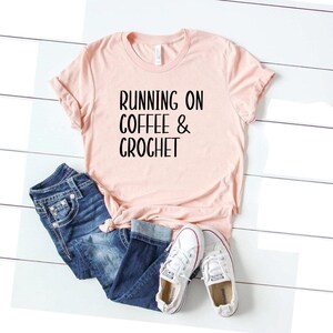 May include: A pink t-shirt with the text "RUNNING ON COFFEE & CROCHET" in black lettering. The shirt is laid out on a white wooden surface with a pair of white sneakers and blue jeans.