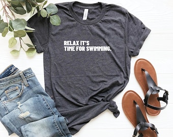 Może przedstawiać: Ciemnoszara koszulka z białym napisem "RELAX IT'S TIME FOR SWIMMING." Koszulka jest zestawiona z niebieskimi dżinsami i brązowymi sandałami z czarnymi paskami. Gałązka zieleni jest widoczna w tle.