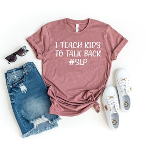 Op de afbeelding: Een mauvekleurig T-shirt met de tekst "I TEACH KIDS TO TALK BACK #SLP" wordt getoond met een spijkerbroek, zonnebril en witte sneakers. Het T-shirt is aan de onderkant geknoopt. De zonnebril is zwart. De sneakers hebben gouden accenten.