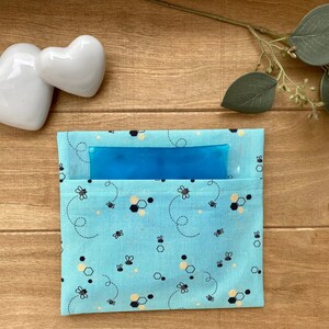 Könnte beinhalten: Eine blaue Stofftasche mit einem Bienen- und Wabenmuster. Die Tasche hat ein transparentes Plastikfenster und enthält einen blauen Eisbeutel.