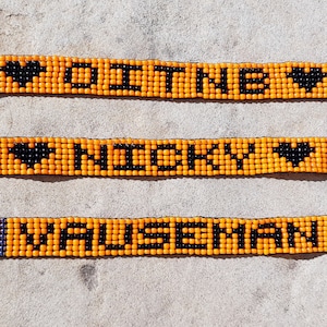 May include: Three orange beaded bracelets with black beads spelling out the names "DITNE", "NICKY", and "HAUSEMAN" with a black heart on each side of the name.